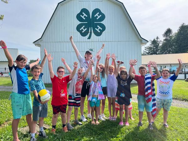 Group of children outdoors in front of a barn with a four-leaf clover symbol, raising hands and smiling.