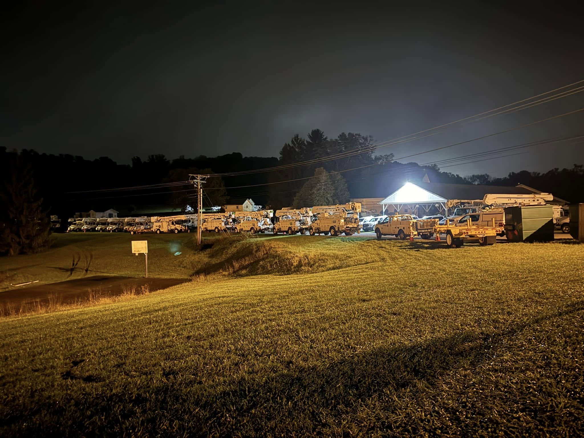 Night scene with a row of vehicles and buildings illuminated, grassy foreground, dark sky, and power lines, no people visible.