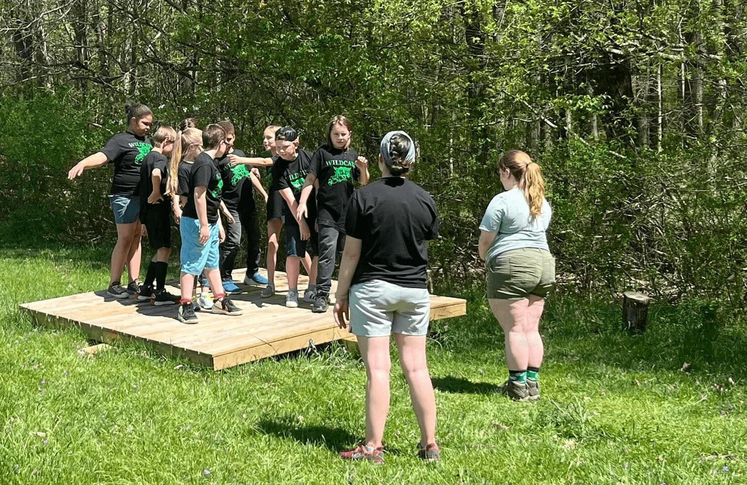 Group of children and adults standing on a wooden platform outdoors in a grassy area with trees, some children wearing black shirts.