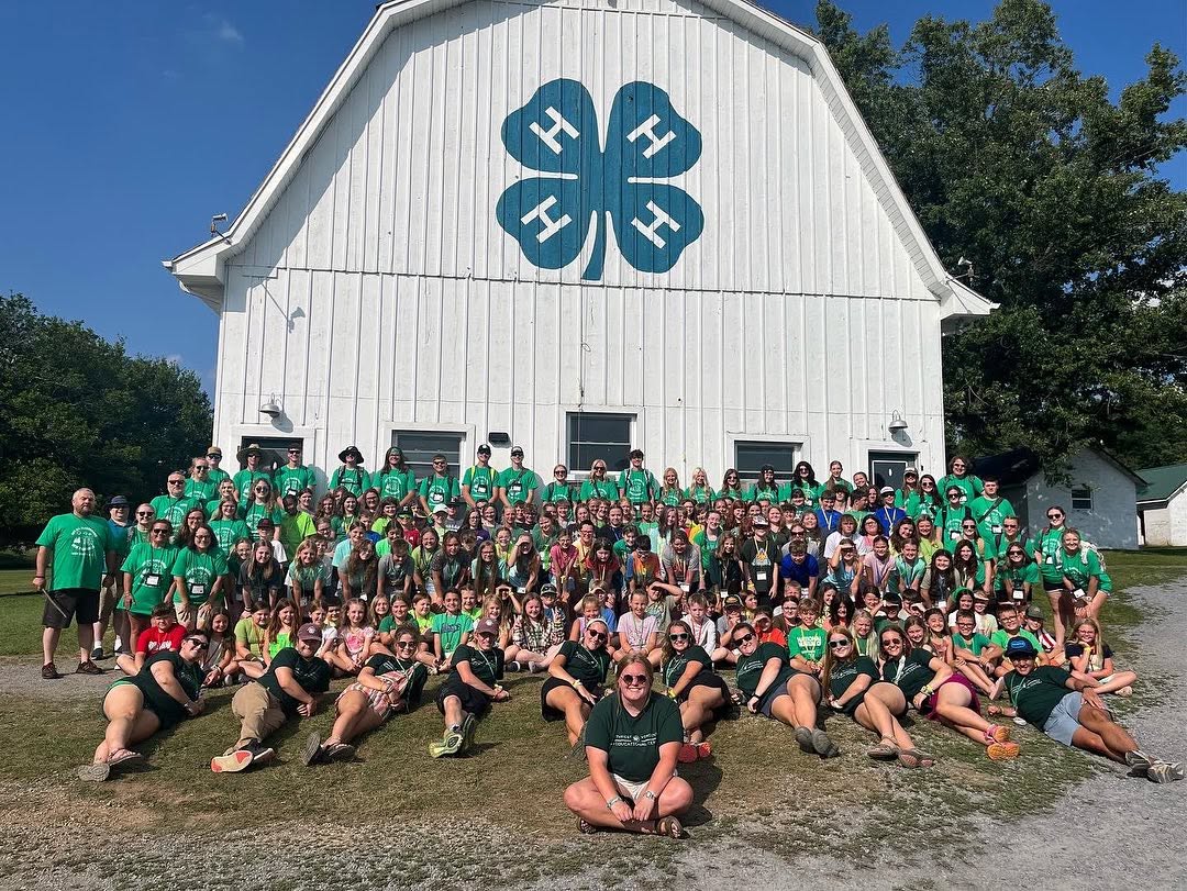 Large group of people gathered outside a white barn with a four-leaf clover logo, some wearing green, on grass with trees in background.
