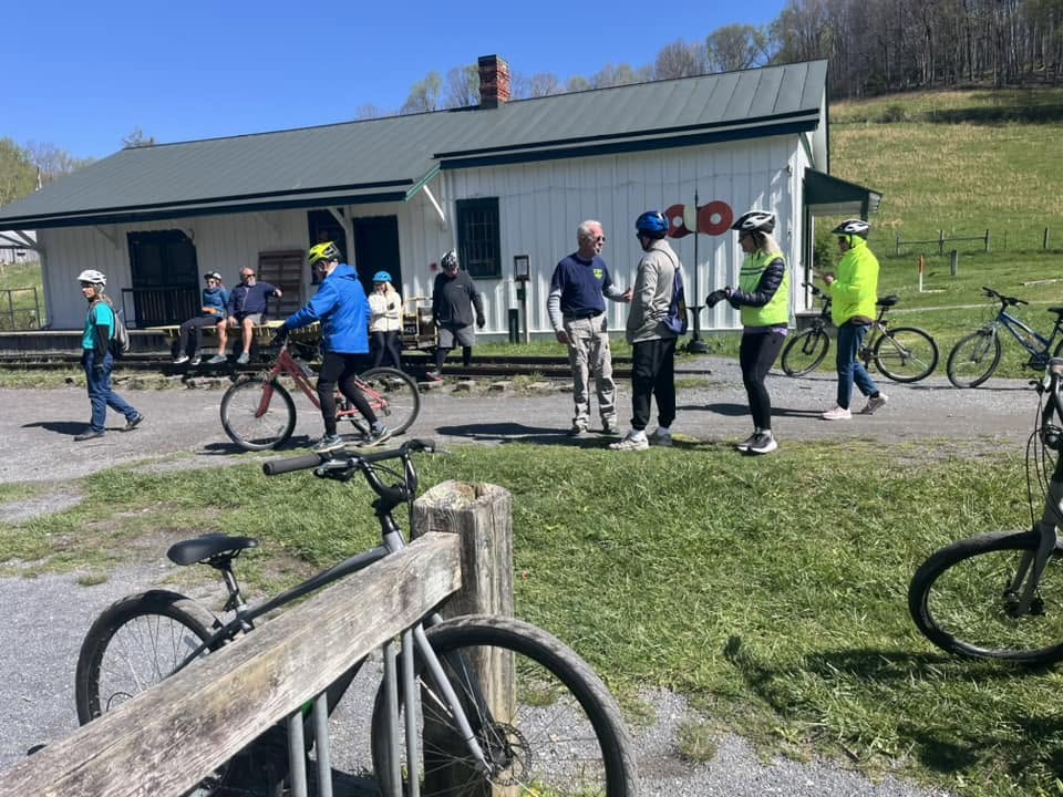 Group of people standing outside a light blue building with bicycles nearby, on a sunny day with green grass and trees.
