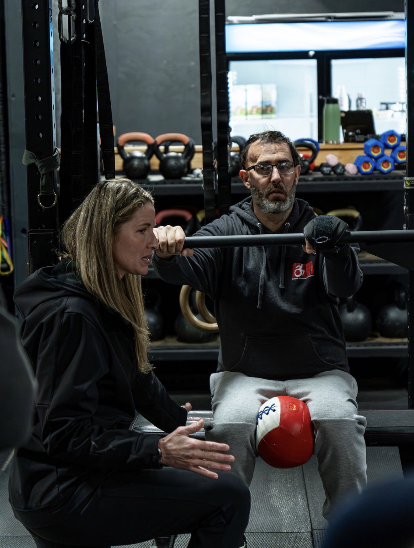 A woman and a man are in a gym, with the woman holding a barbell and the man coaching her, with gym equipment in the background.