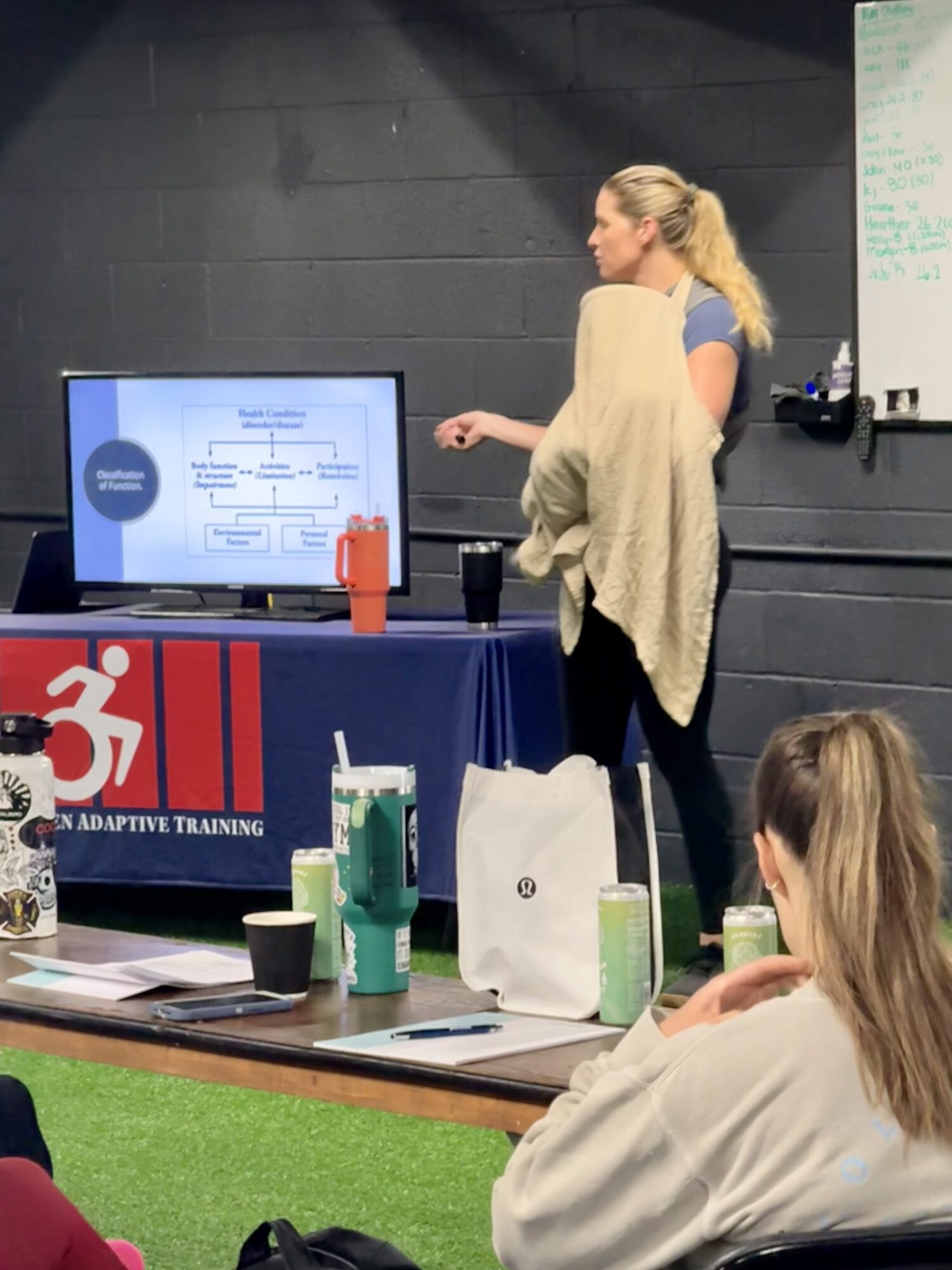 Two women stand near a presentation screen in a classroom, with a table and various items in the foreground.