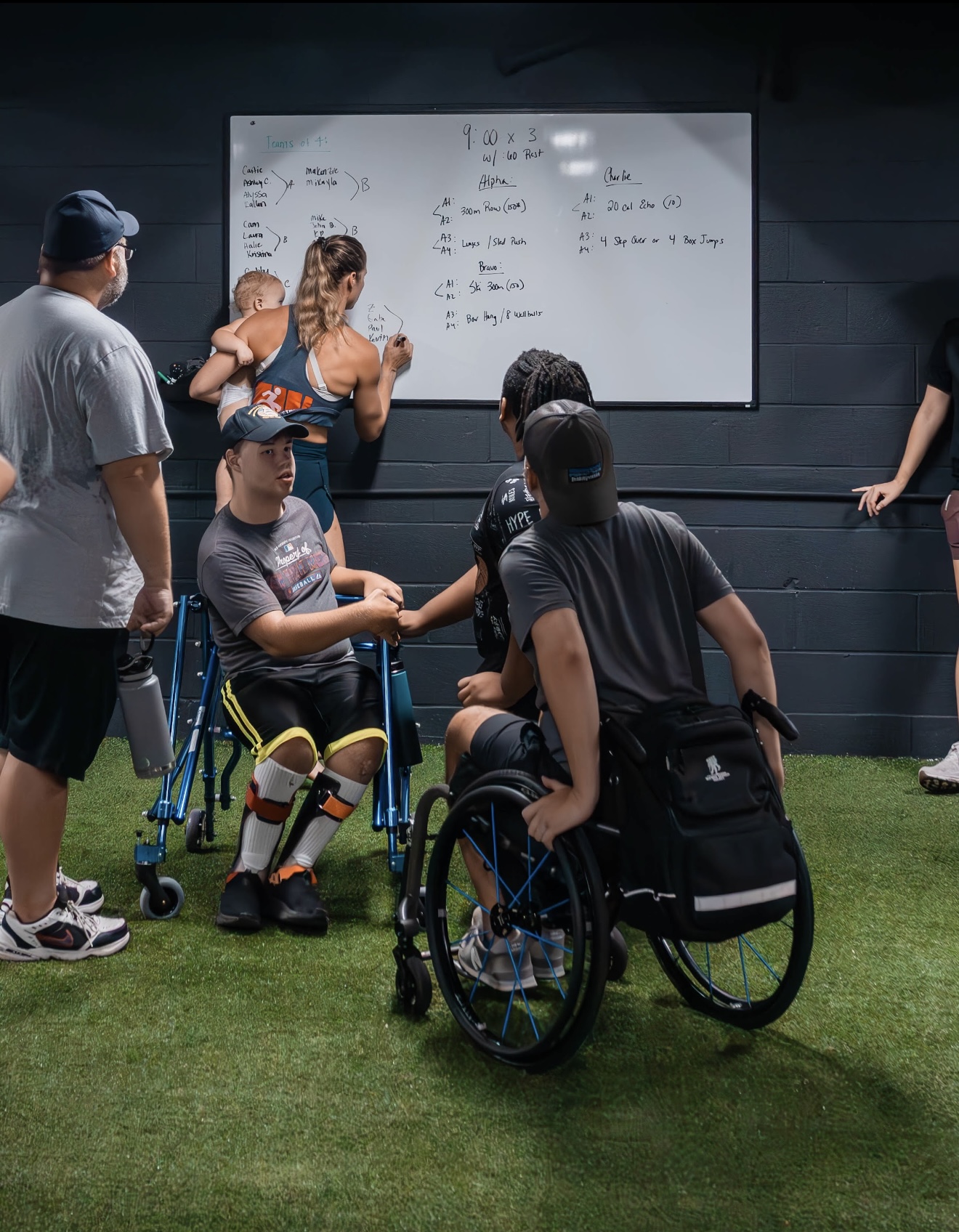 Group of people, including a person in a wheelchair, gathered in front of a whiteboard with writing, in a room with dark walls and green floor.