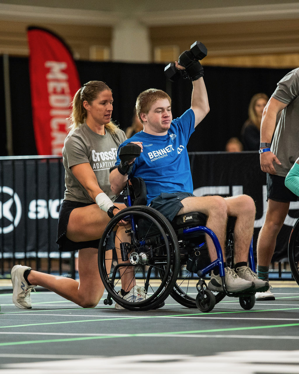 A person in a wheelchair lifts a dumbbell while a woman kneels beside them, in an indoor sports setting.