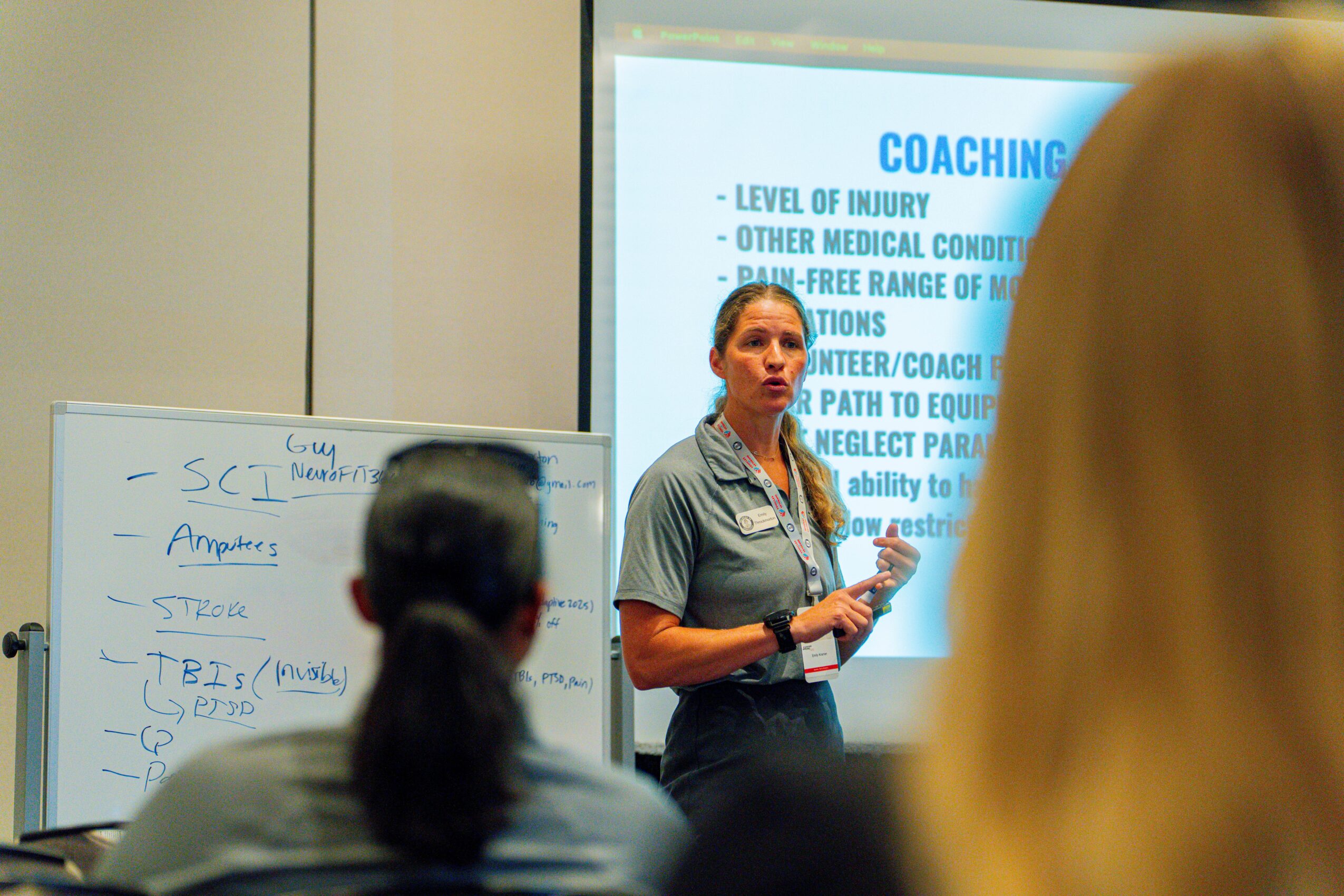 Woman giving a presentation in a classroom with a projected slide and a whiteboard, audience members visible in foreground.