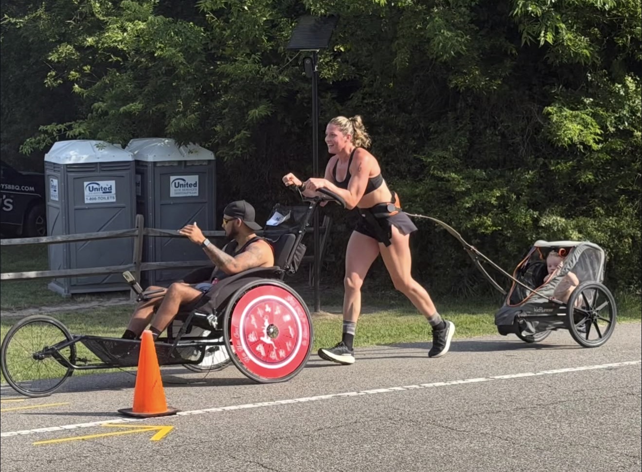 Person in wheelchair being pulled by another person on a leash, with a child in a stroller nearby, on a road with trees.