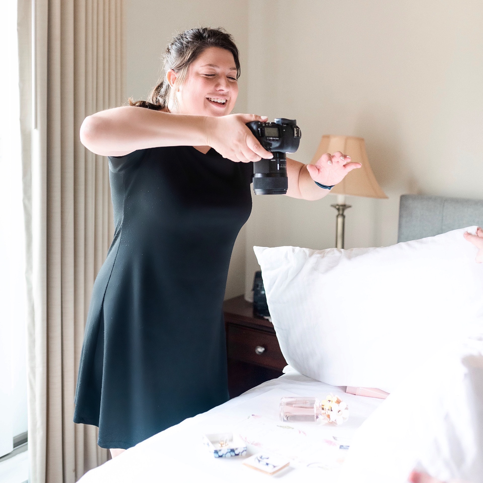 Woman smiling and taking a photo with a camera in a bedroom, with a bed and bedside lamp visible.