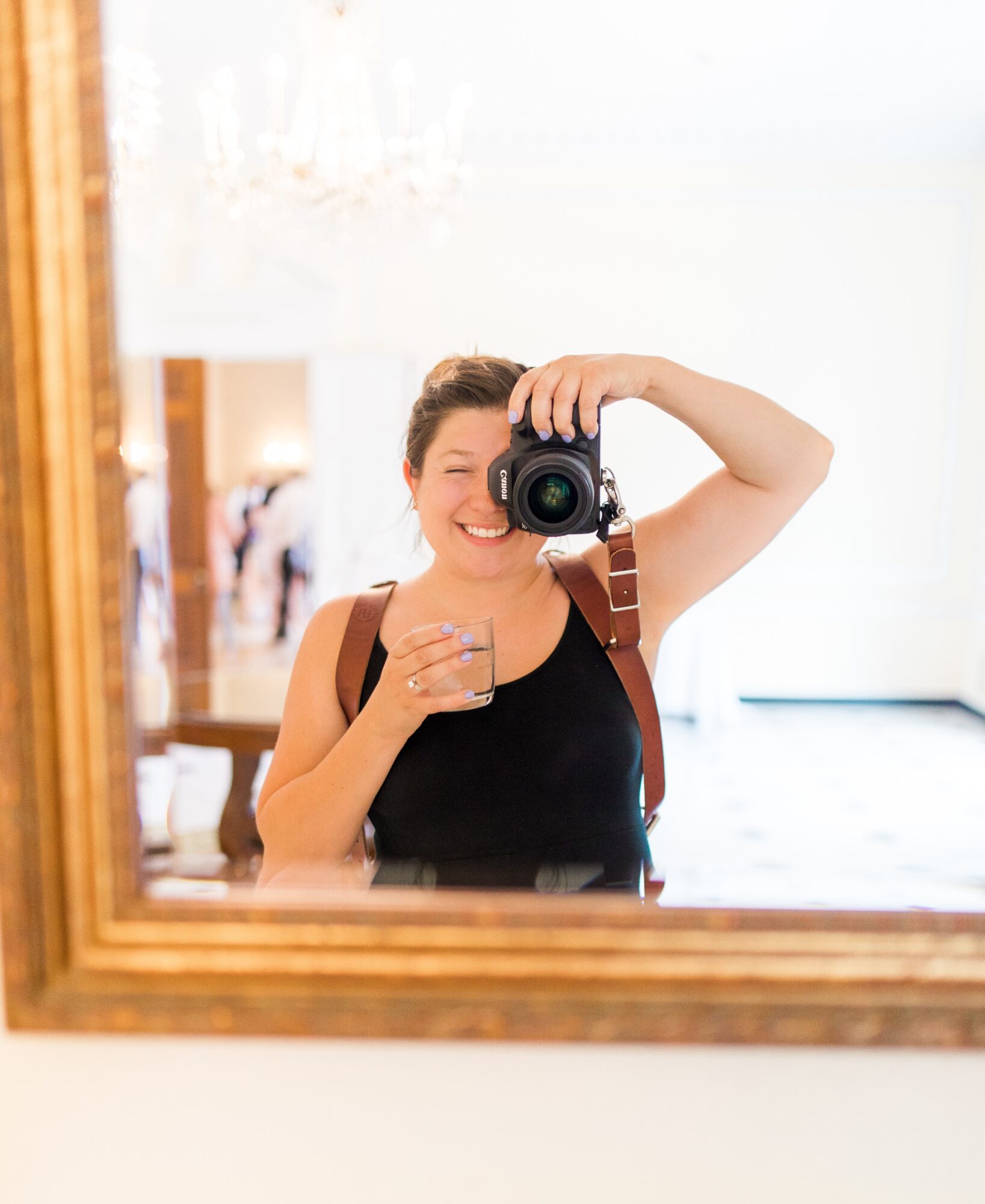 Person taking a selfie in a mirror, holding a camera with one hand and a drink with the other, smiling, indoors.