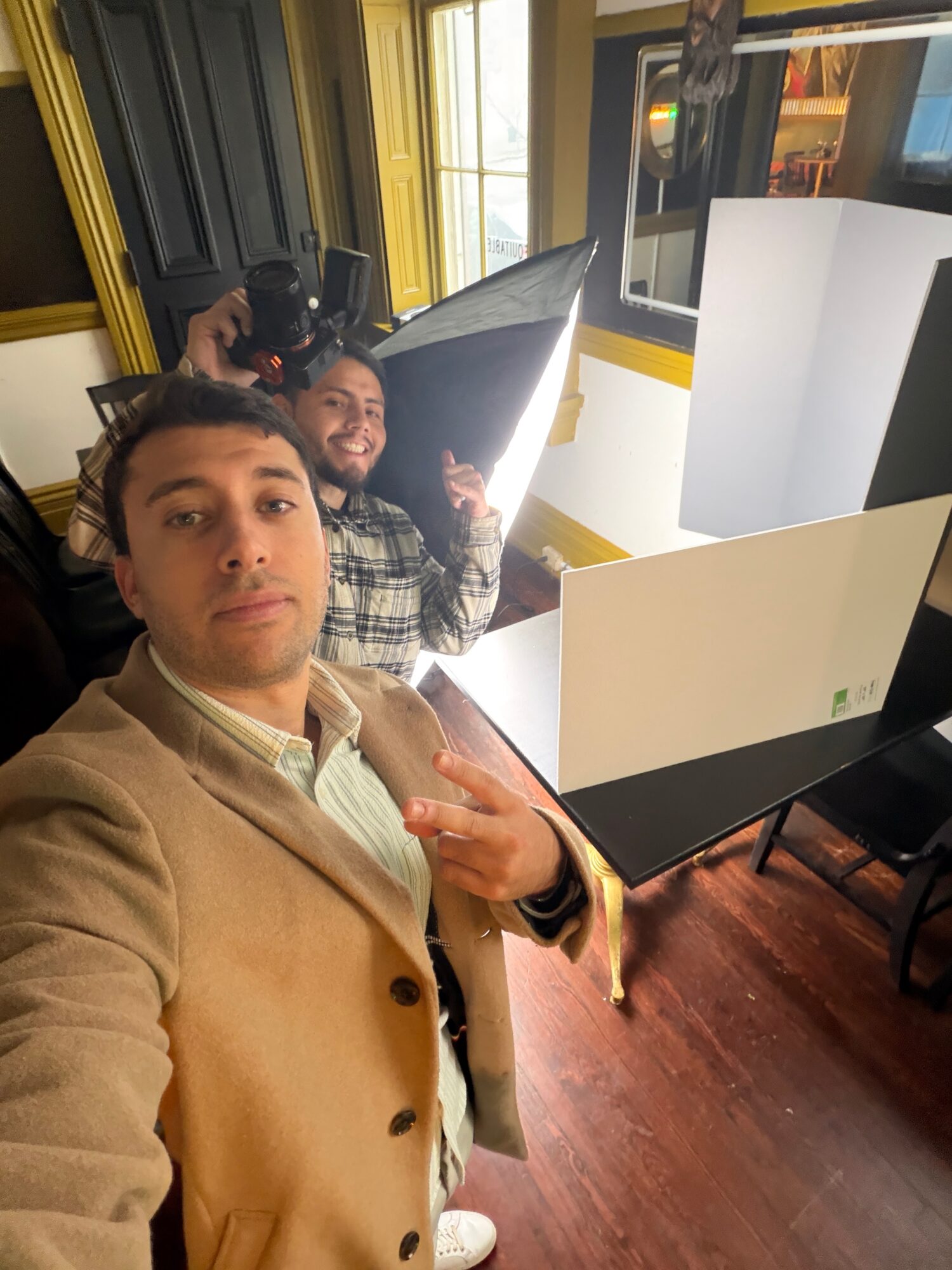 Two men taking a selfie in a room with large windows, desks, and computer monitors, one making a peace sign.