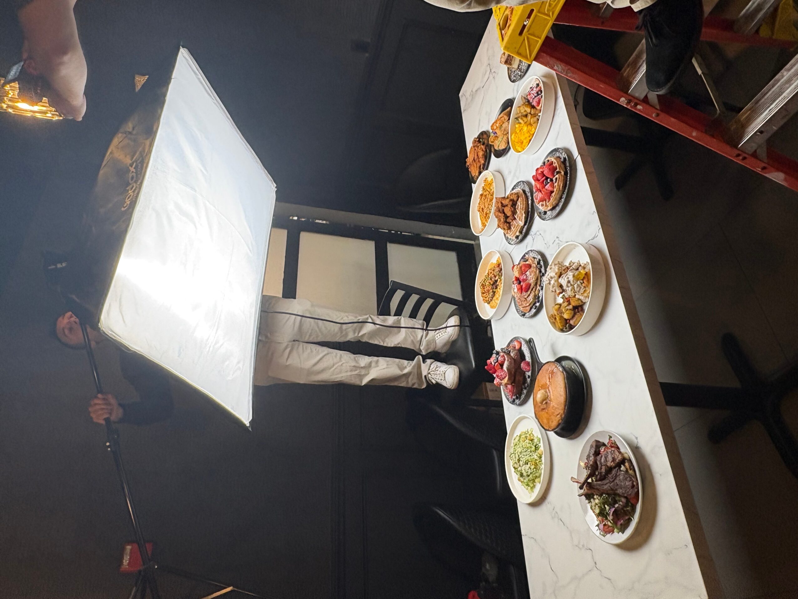 Table with various dishes and drinks, person standing behind, studio lighting setup, black background.