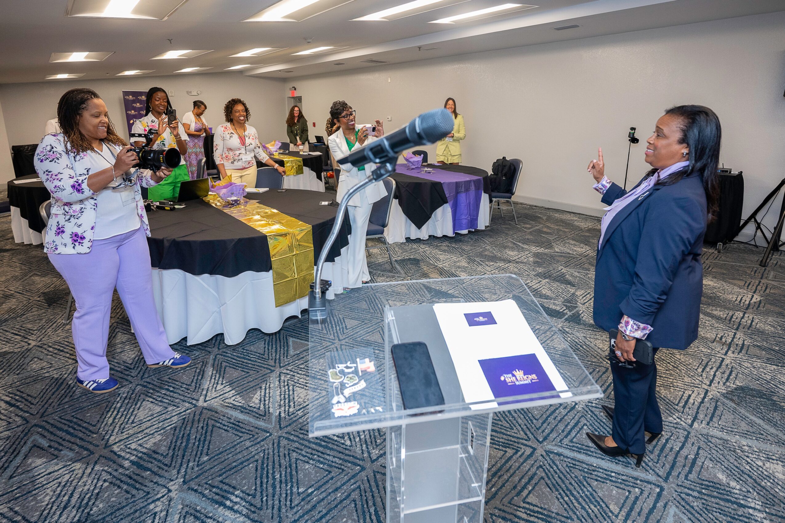 Group of women in a room with a robotic arm, some taking photos, others observing, tables with decorations, woman speaking.