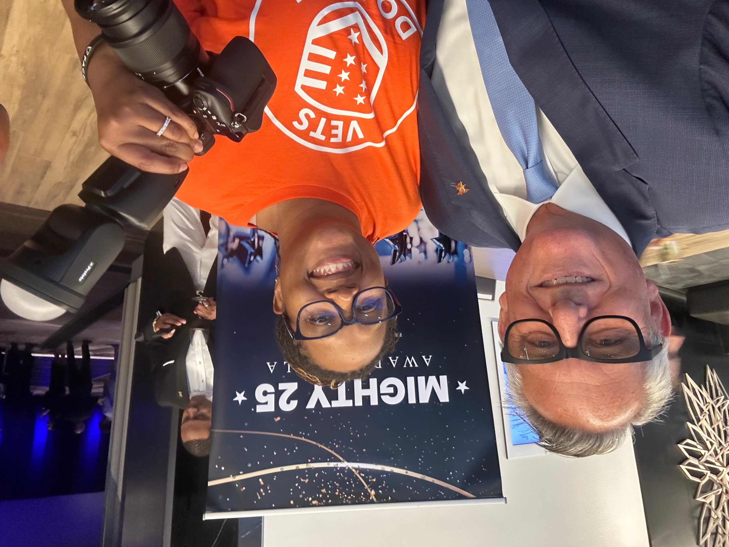 Two people smiling, one holding a camera, standing in front of a sign that reads 'Midcity 25'.