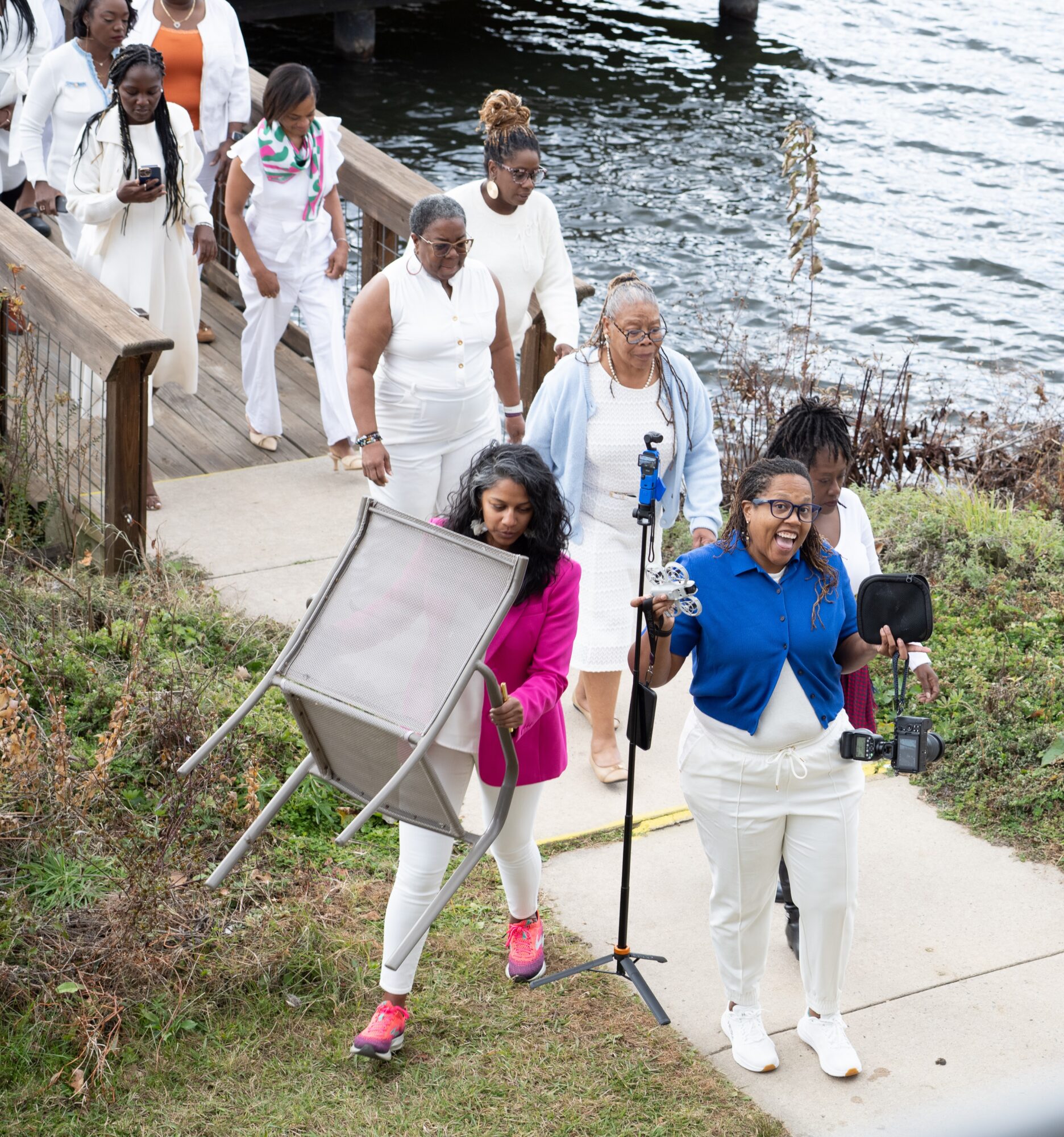 Group of people walking along a waterfront path, some carrying bags, with water and rocks in the background.