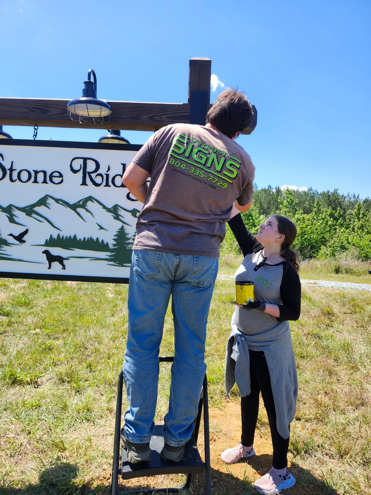 Two people standing outdoors near a sign with trees and a crocodile illustration, under a clear blue sky.