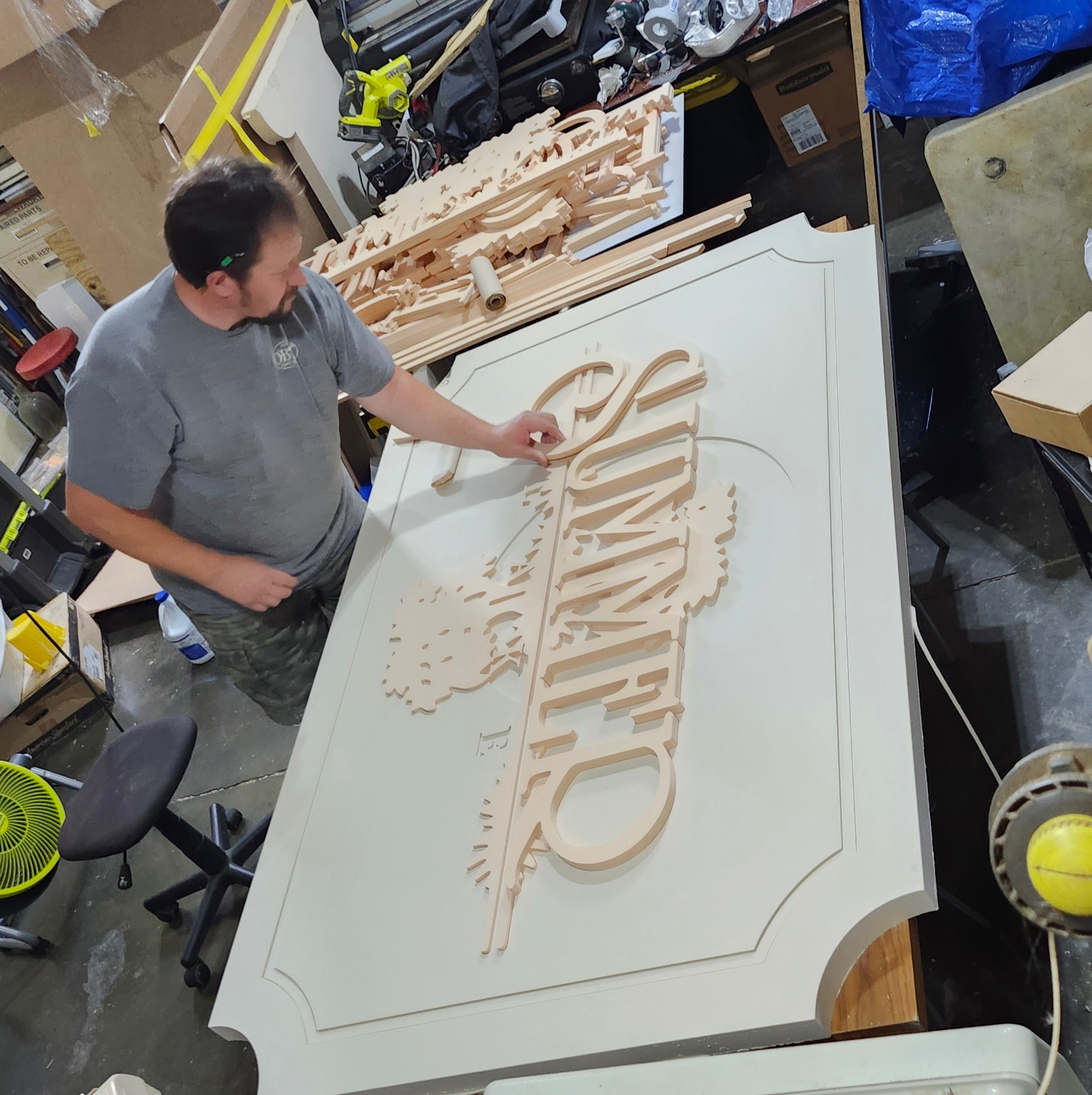 Person working on a large white sign with carved text, surrounded by tools and materials in a workshop.