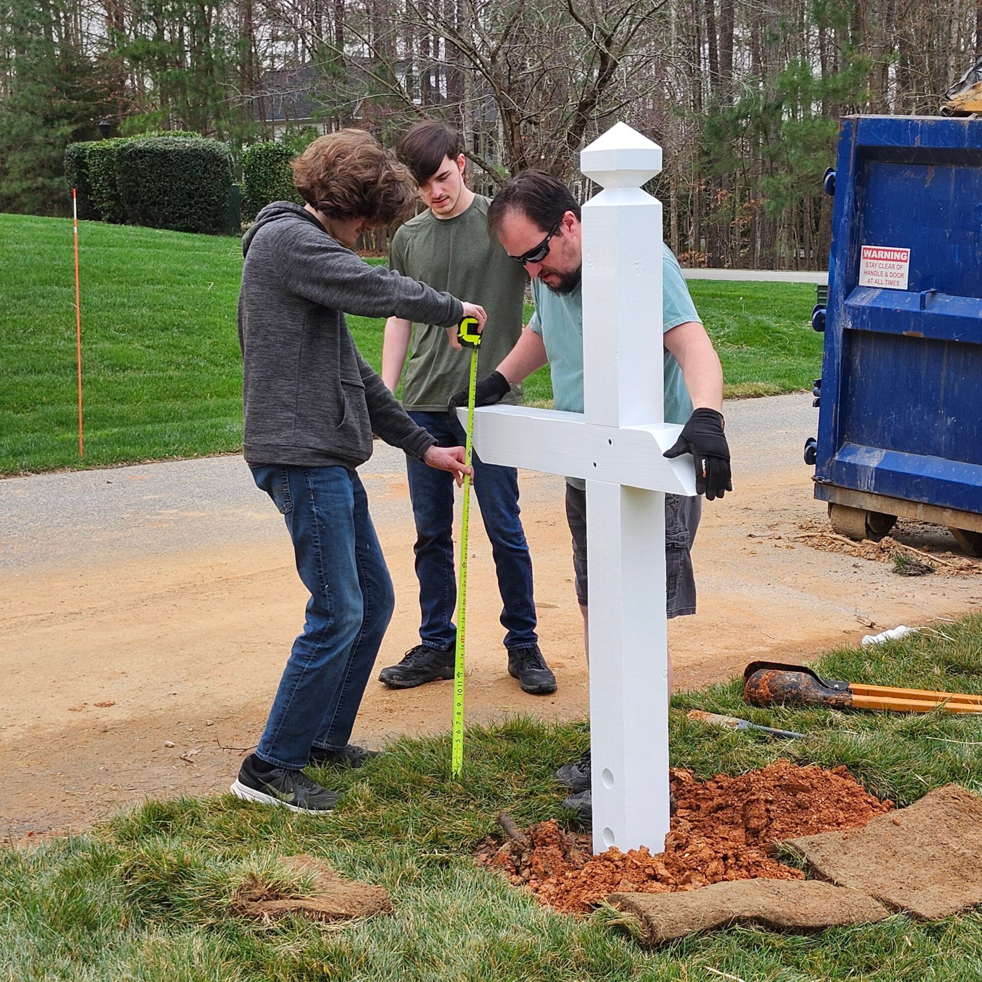 Two people installing a white post into the ground outdoors, with tools and a blue container nearby.