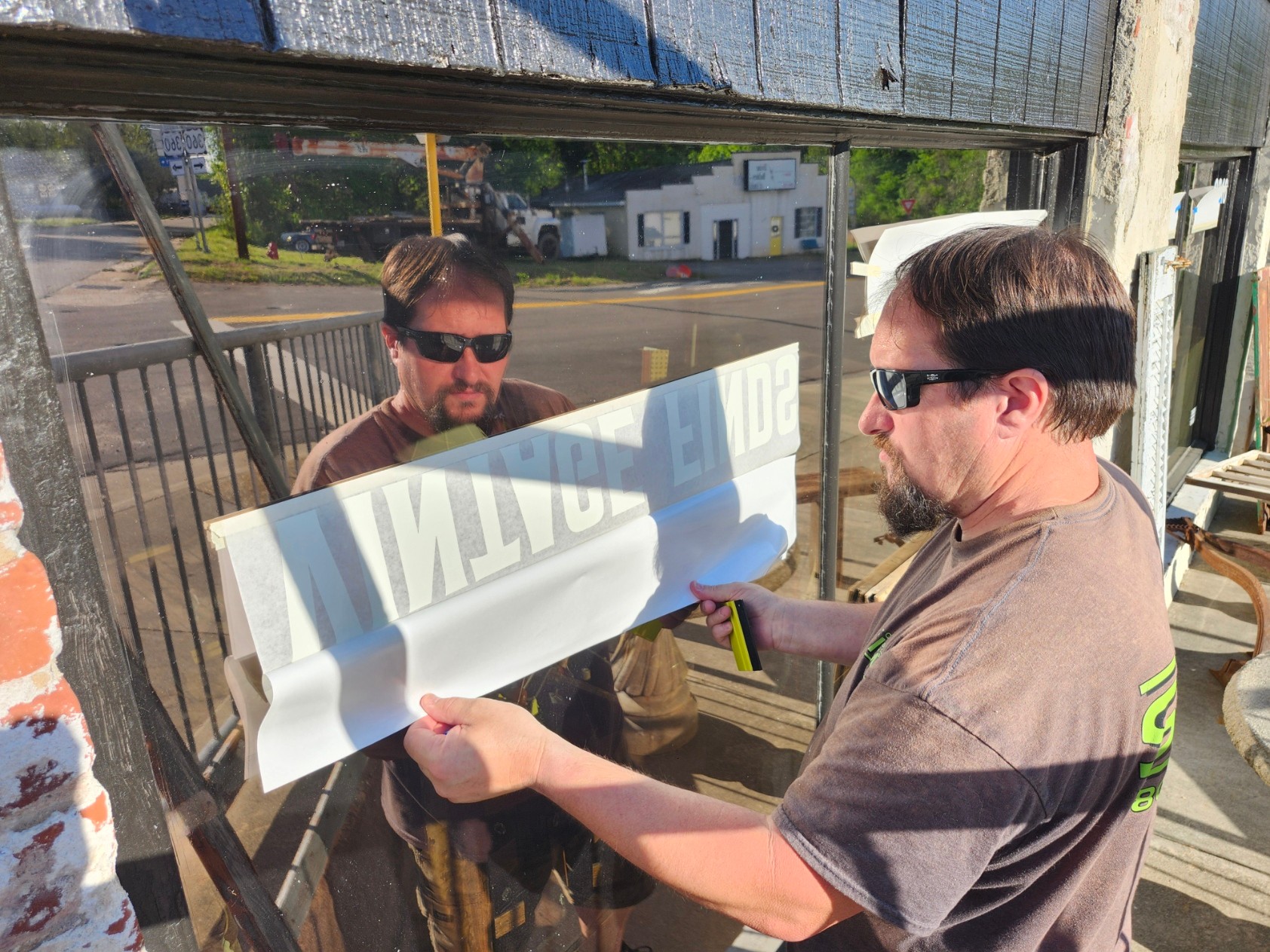 Two men wearing sunglasses are holding a large white object outdoors, with a building and trees in the background.