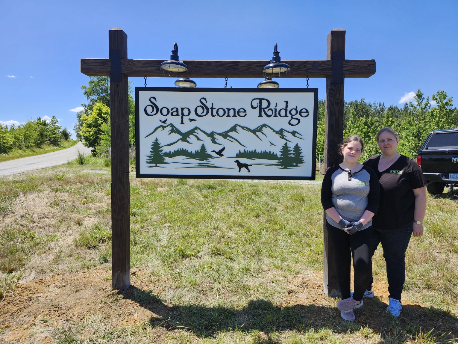 Two women stand next to a sign that reads 'Soap Stone Ridge' with mountains and a dog silhouette, outdoors on a sunny day.