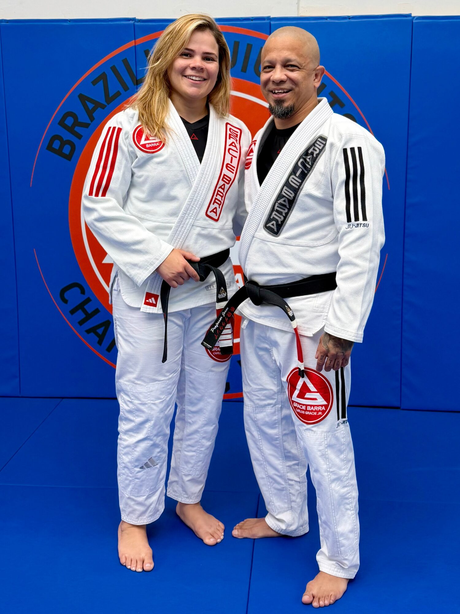Two people in Brazilian Jiu-Jitsu gis standing on a blue mat, smiling, with a Brazilian Jiu-Jitsu logo in the background.