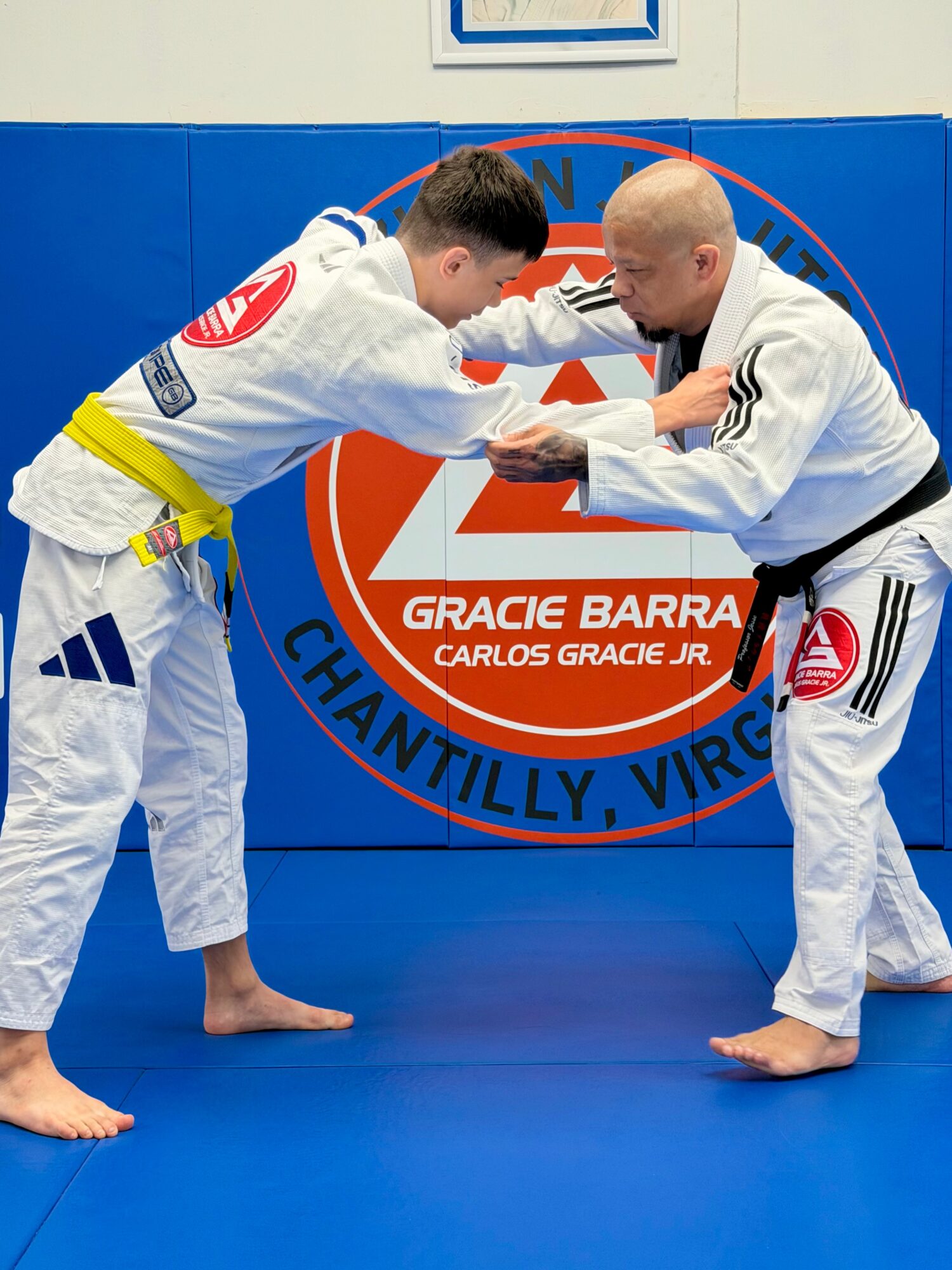 Two people in Brazilian Jiu-Jitsu uniforms practicing a technique on a blue mat, with a logo in the background.