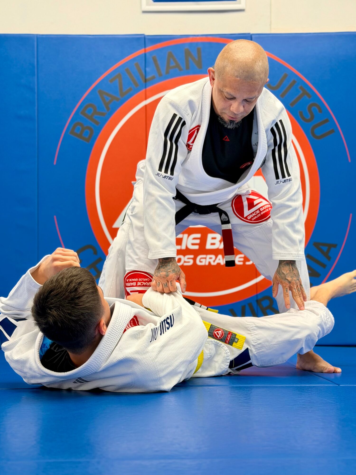 Two people practicing Brazilian Jiu-Jitsu on a blue mat, one person on top and the other on the ground, with a circular logo in the background.