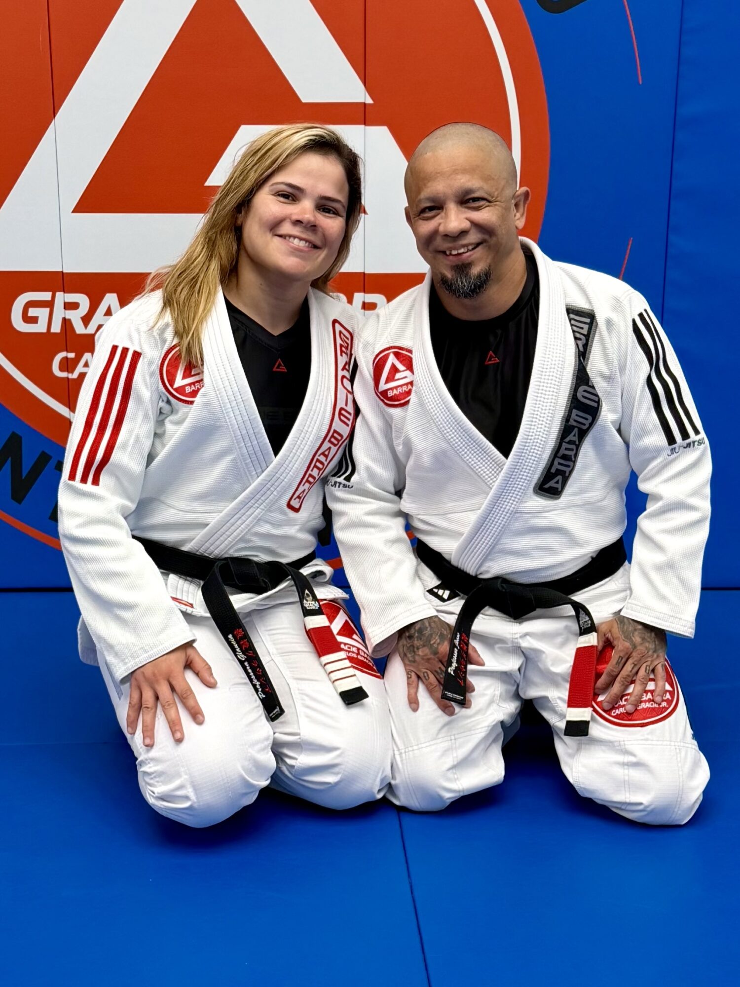 Two people in Brazilian Jiu-Jitsu gis kneeling on a blue mat, smiling, with a large red and white logo behind them.