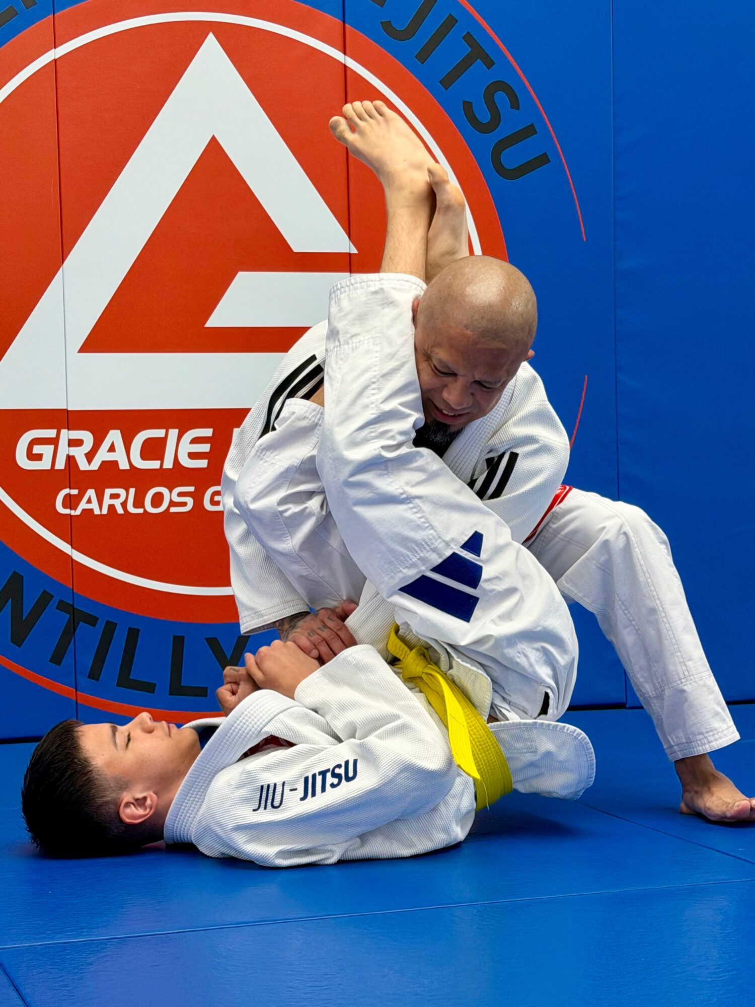 Two people practicing Brazilian Jiu-Jitsu on blue mat, one in white gi with yellow belt, the other in white gi, near a red and blue wall with logo.
