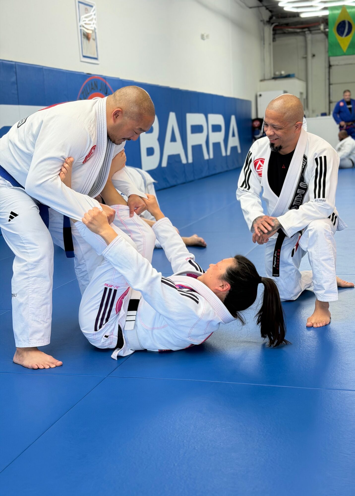 Three people in Brazilian Jiu-Jitsu gis practicing on blue mat, one person on the ground, two standing, in a training facility.