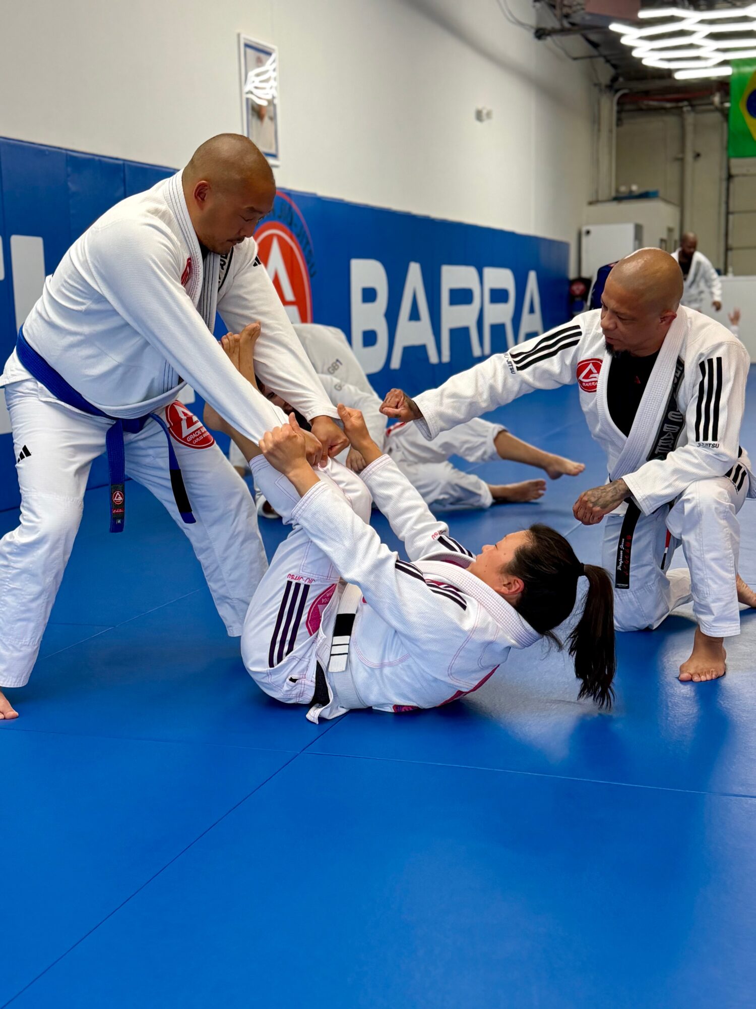 Two men in gi uniforms are practicing Brazilian Jiu-Jitsu on a blue mat, with one woman on the ground and others assisting.