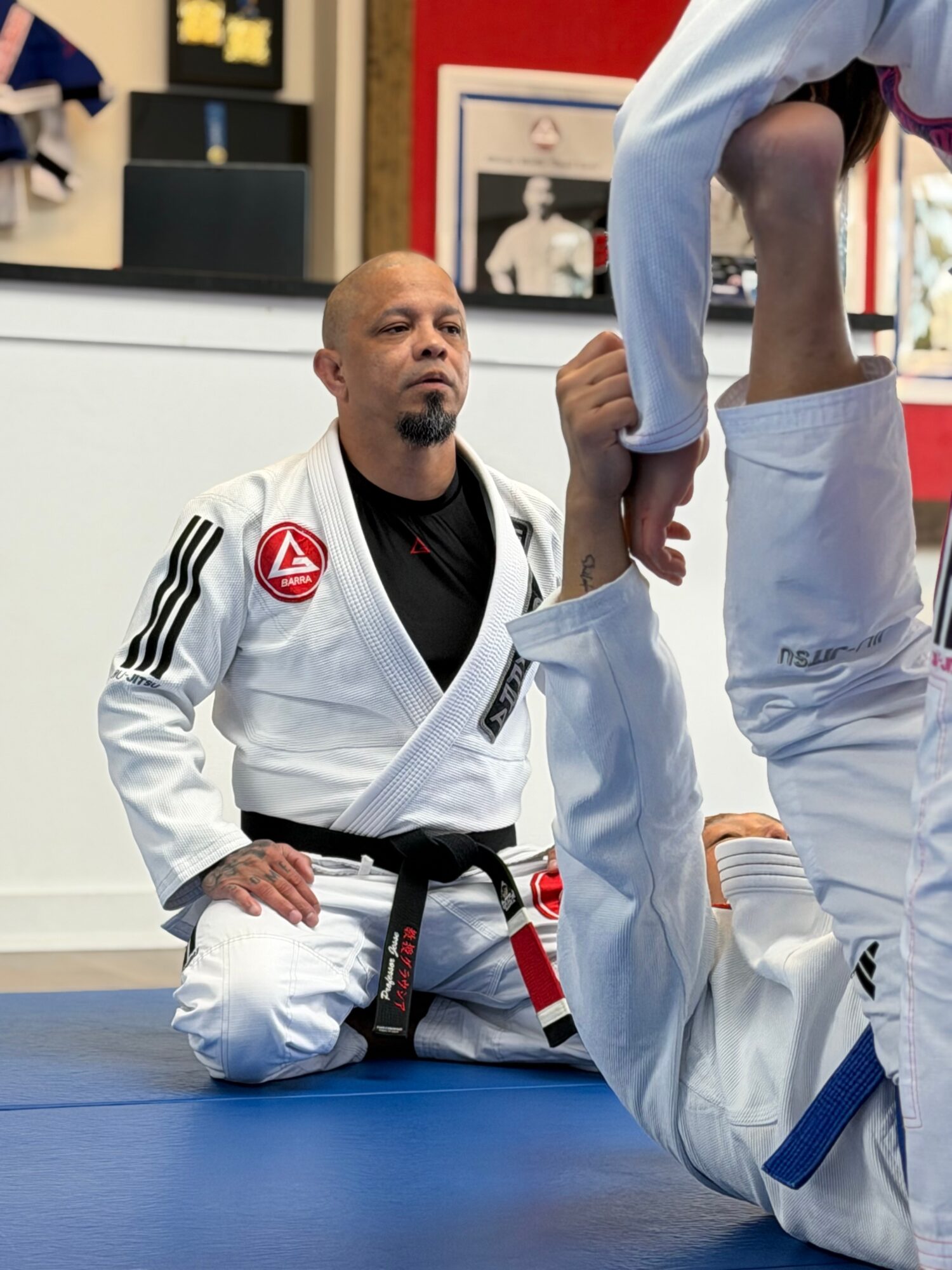 Martial arts instructor kneeling on blue mat, observing student practicing Brazilian Jiu-Jitsu, in a training facility with posters and equipment.