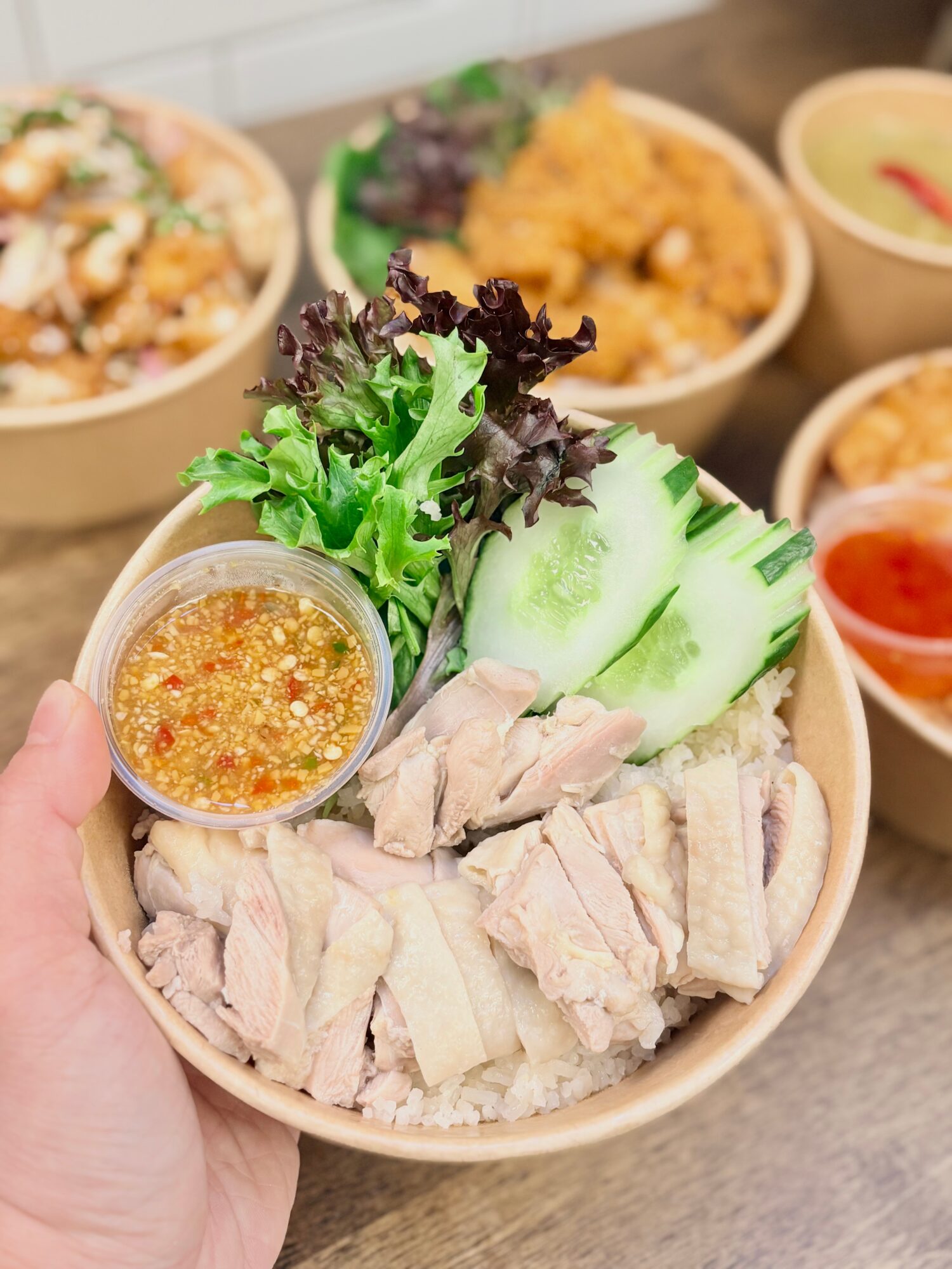 Bowl with sliced chicken, cucumber, lettuce, and a small container of dipping sauce, with other bowls of food in background.