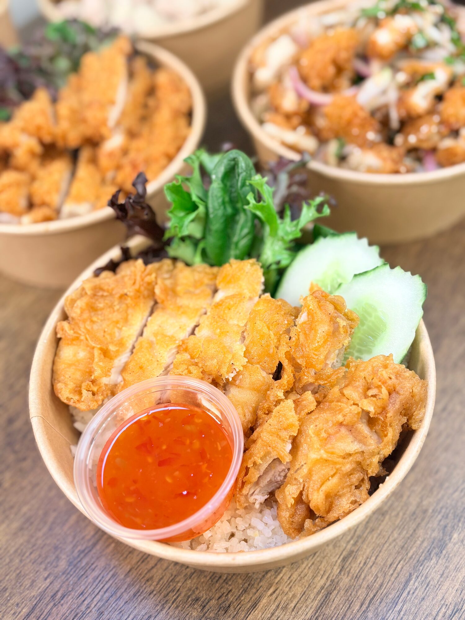 Bowl with fried chicken pieces, rice, cucumber slices, lettuce, and a small container of red dipping sauce.