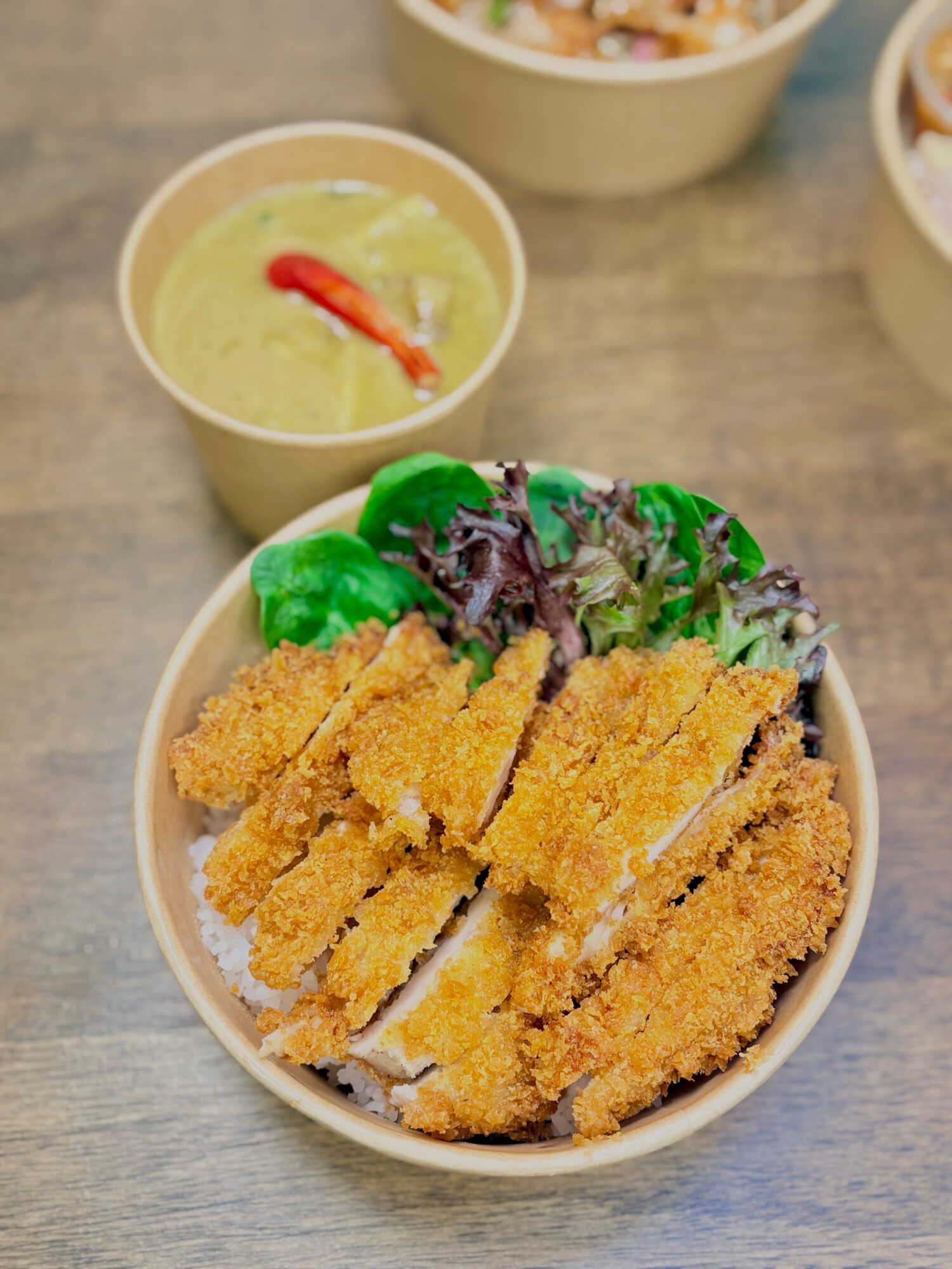 Bowl of fried chicken strips with green vegetables, accompanied by a cup of green dipping sauce with a red chili, on a wooden surface.