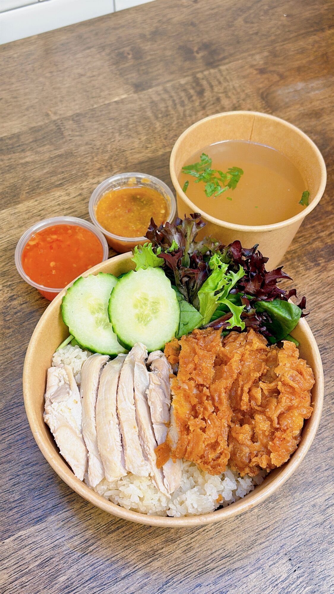 Bowl with sliced cucumber, fried chicken, shredded chicken, lettuce, and sauces, with a cup of soup on a wooden table.