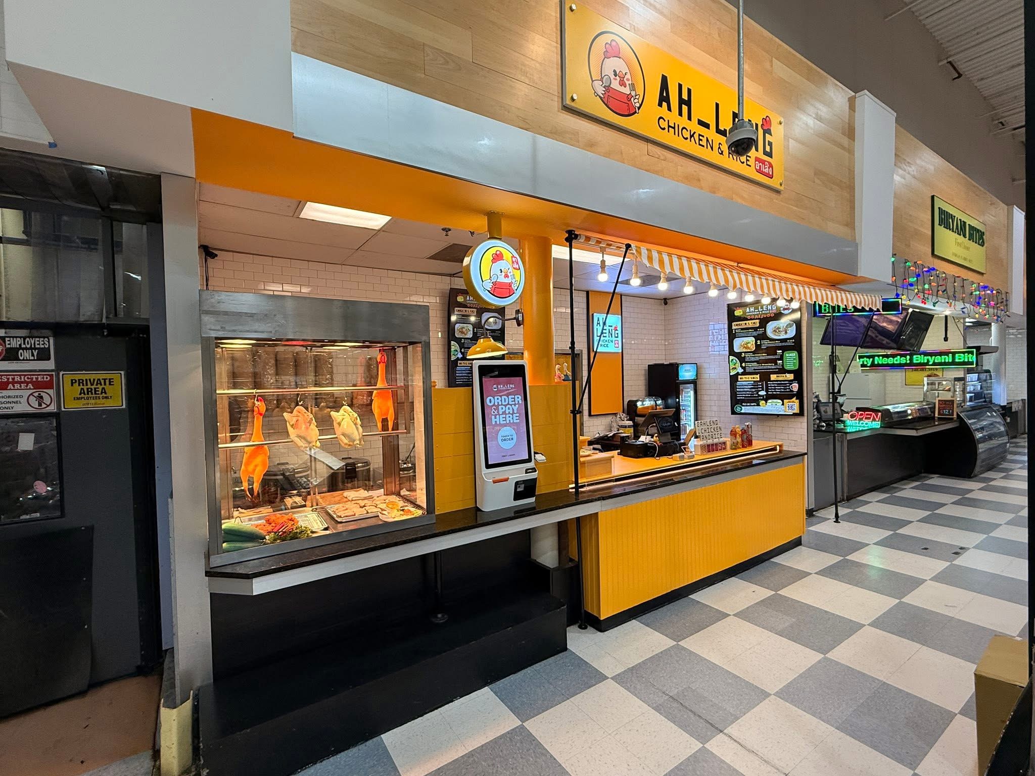 Food counter with a display case, digital menu, and a yellow and orange sign above, in a shopping mall.