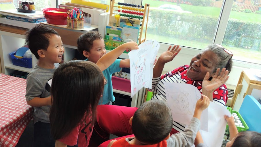 A teacher interacts with five children in a classroom near a window, smiling and raising hands, with educational materials nearby.