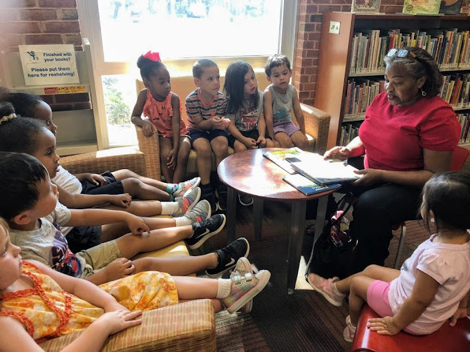 Group of children sitting on a bench and chairs listening to a woman reading a book in a library.