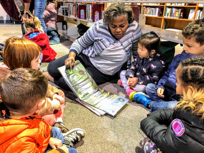 Adult woman reading a book to children seated on the floor in a library, surrounded by bookshelves.