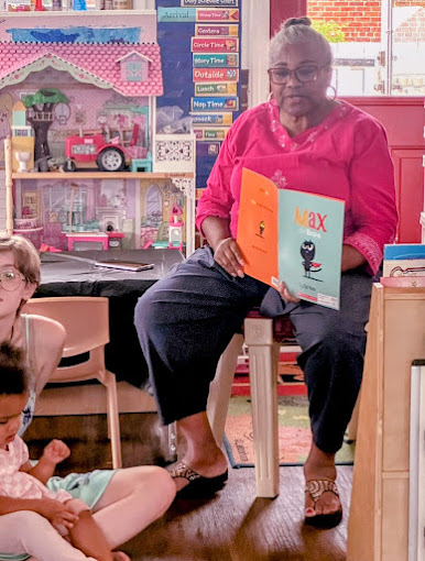Woman sitting on a chair reading a children's book in a colorful room with toys and books, children nearby, wooden floor.