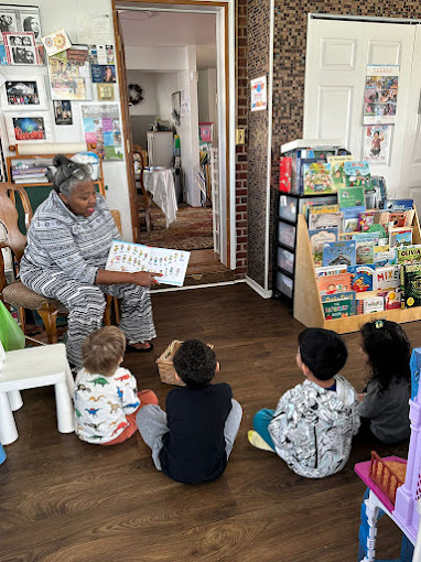 An elderly woman reading a book to four children sitting on the floor in a room with bookshelves and a doorway to another room.