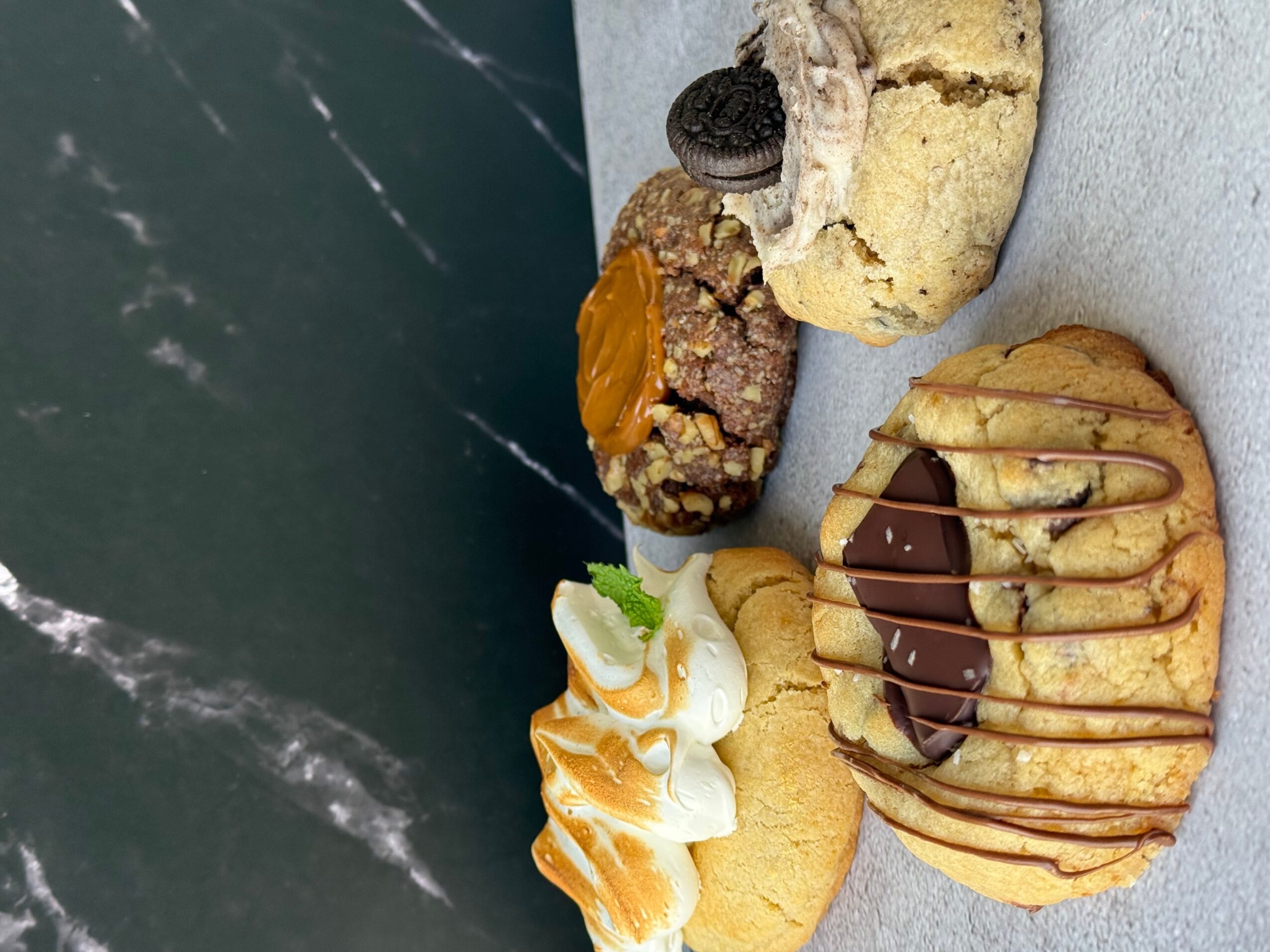Four cookies with various toppings and drizzles on a white surface, with a dark background. Word count: 20.