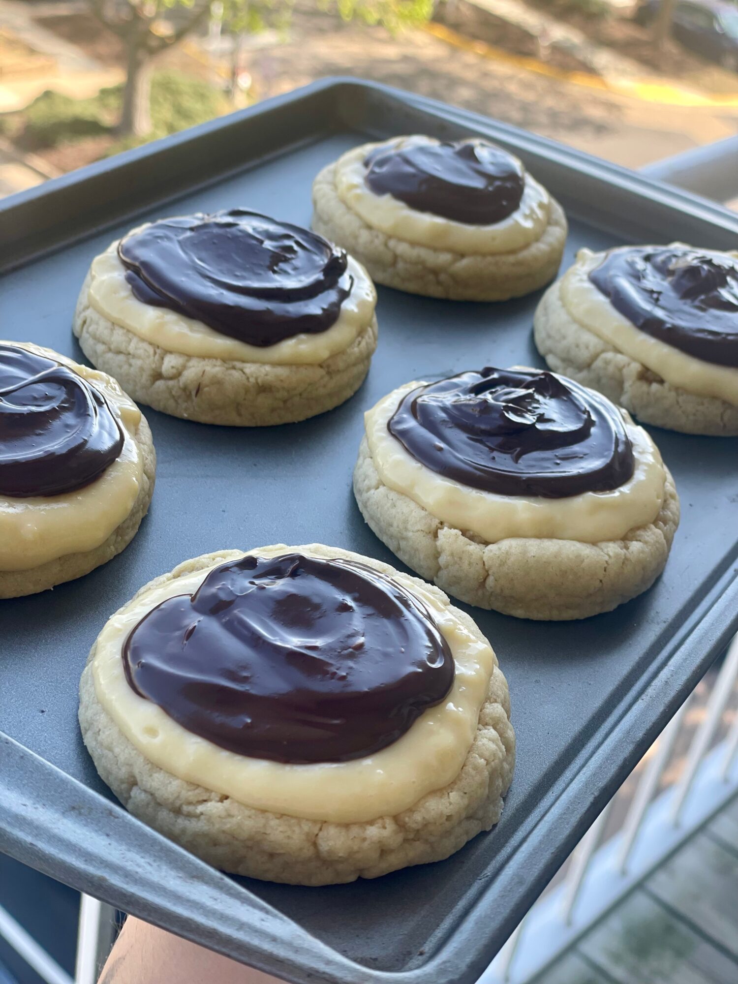 Six cookies with chocolate topping on a tray, outdoors with trees and a building in the background.