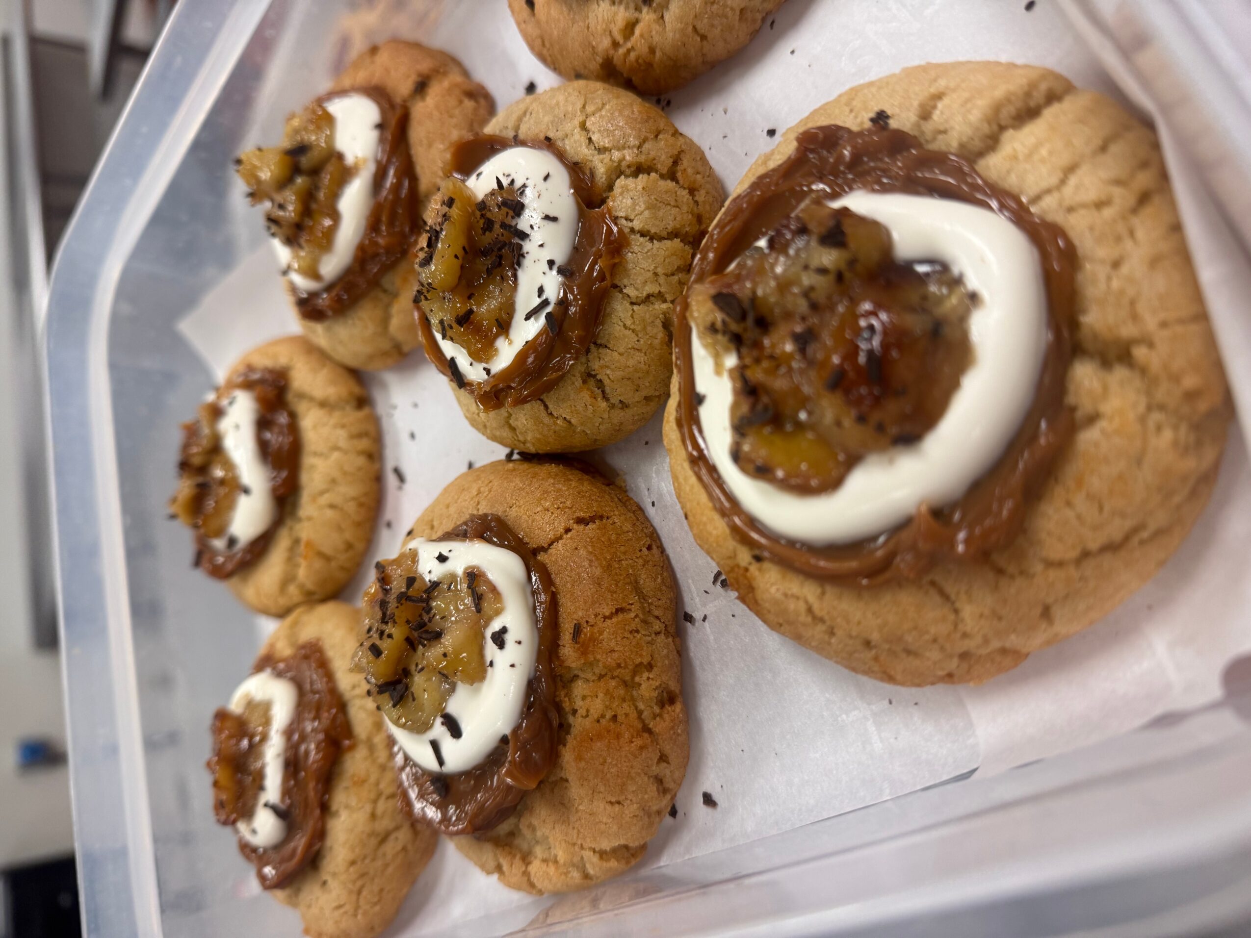Cookies topped with cream, caramel, and candied fruit, arranged in a container on a white paper sheet.