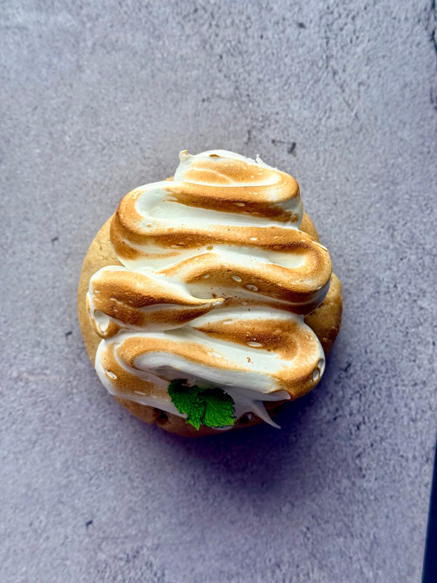 A cupcake with white and caramel-colored frosting topped with a small green leaf, on a gray surface.