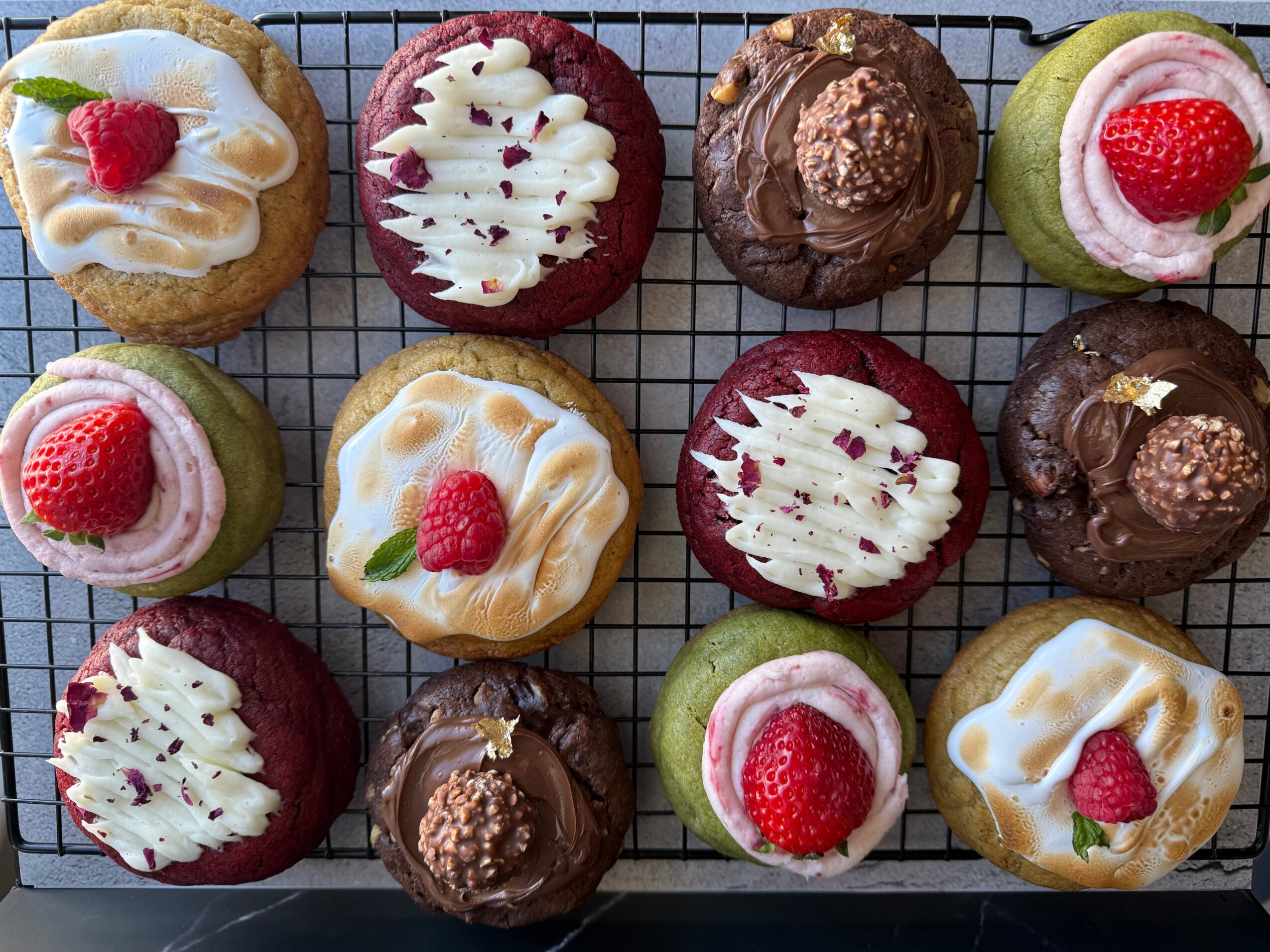 Assorted cupcakes with frosting, strawberries, and toppings on a wire rack, arranged in a grid pattern.