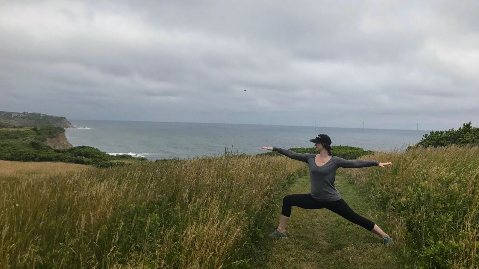 Person practicing yoga outdoors on a grassy path near the coast with arms and legs extended, cloudy sky above.