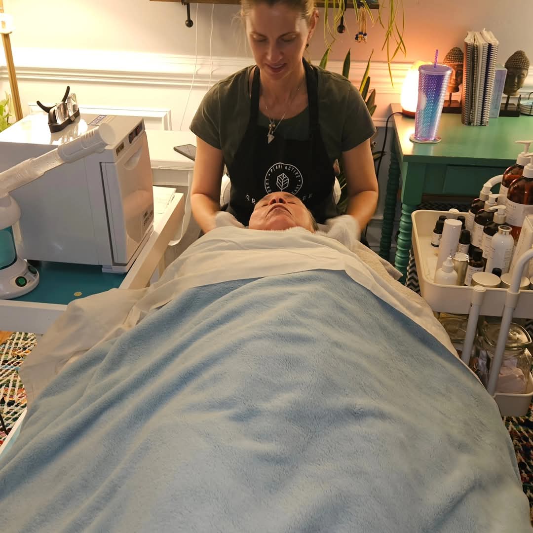 A woman in a black apron leans over a person lying on a bed, in a room with skincare products and equipment.