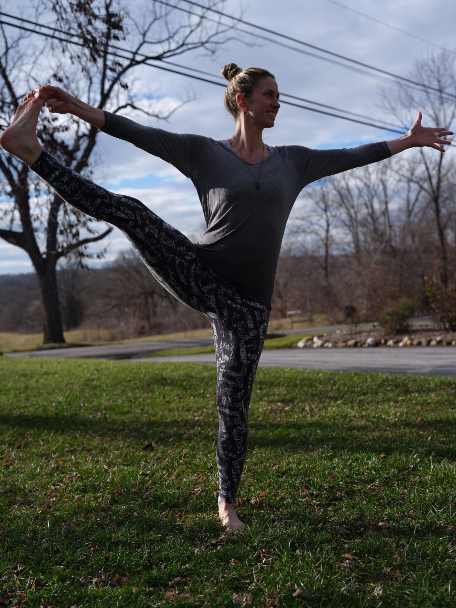 Woman performing a yoga pose outdoors on grass, with trees and cloudy sky in background.