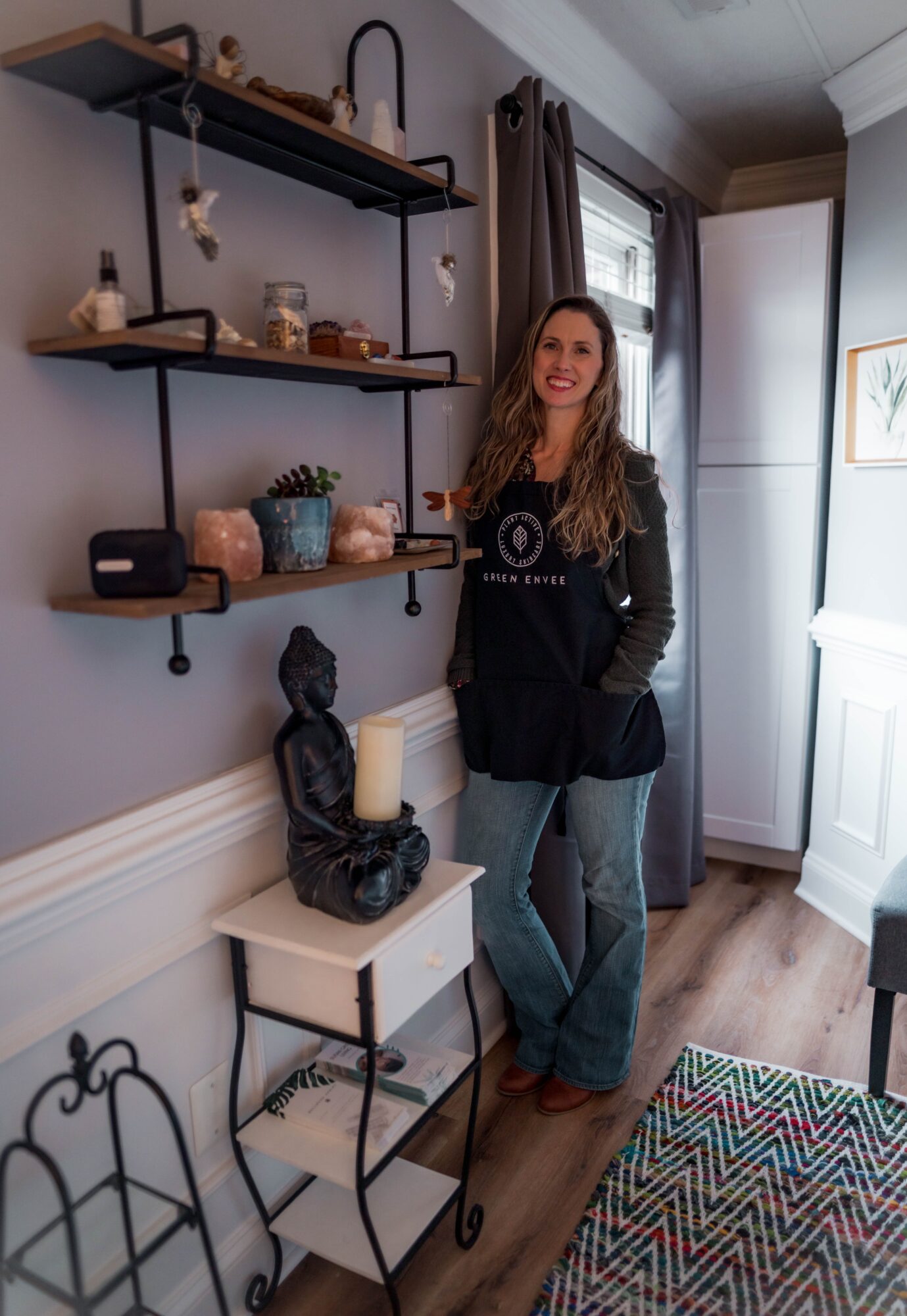 Woman with long hair standing in a room with shelves, a small table, and a patterned rug, smiling at the camera.